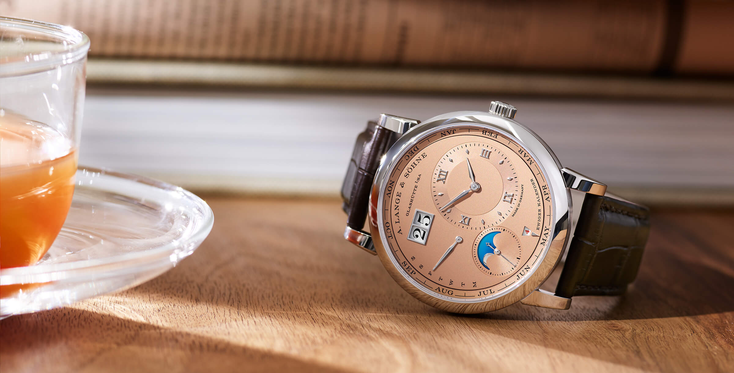 A. Lange & Söhne Perpetual Calendar com mostrador salmão e caixa em ouro branco deitado de lado numa mesa de madeira com livros de fundo e uma chávena de chá