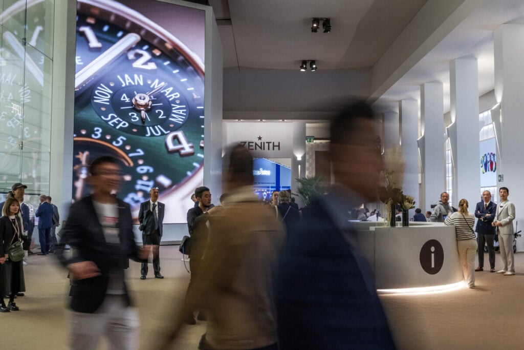 Imagem do interior do Salão Watches and Wonders 2025 com pessoas a passarem e ao fundo com destaque para um écrã com um relógio patek philippe em grande escala