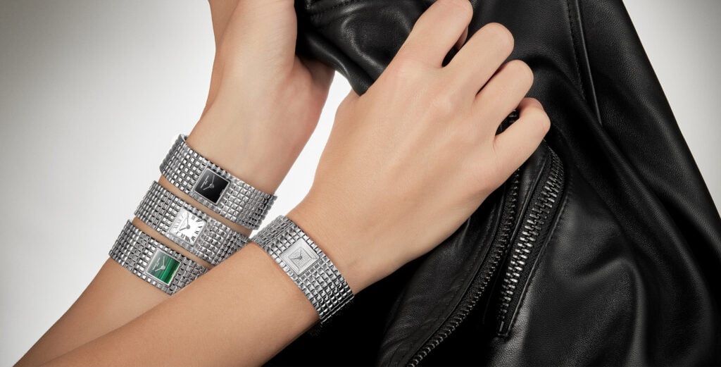 Quatro relógios femininos com braceletes metálicas prateadas estilo pulseira tipo manchette, usados em ambos os pulsos, segurando uma jaqueta de couro preta.