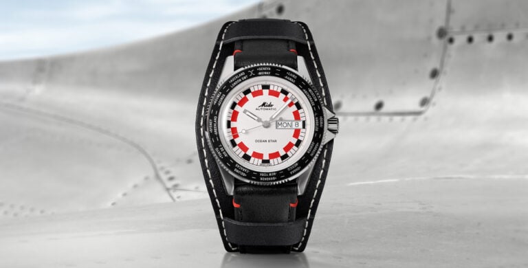 Relógio Mido Ocean Star Worldtimer em fundo metálico, com visual de mostrador branco e detalhes vermelhos, posicionado sobre superfície industrial com céu ao fundo.