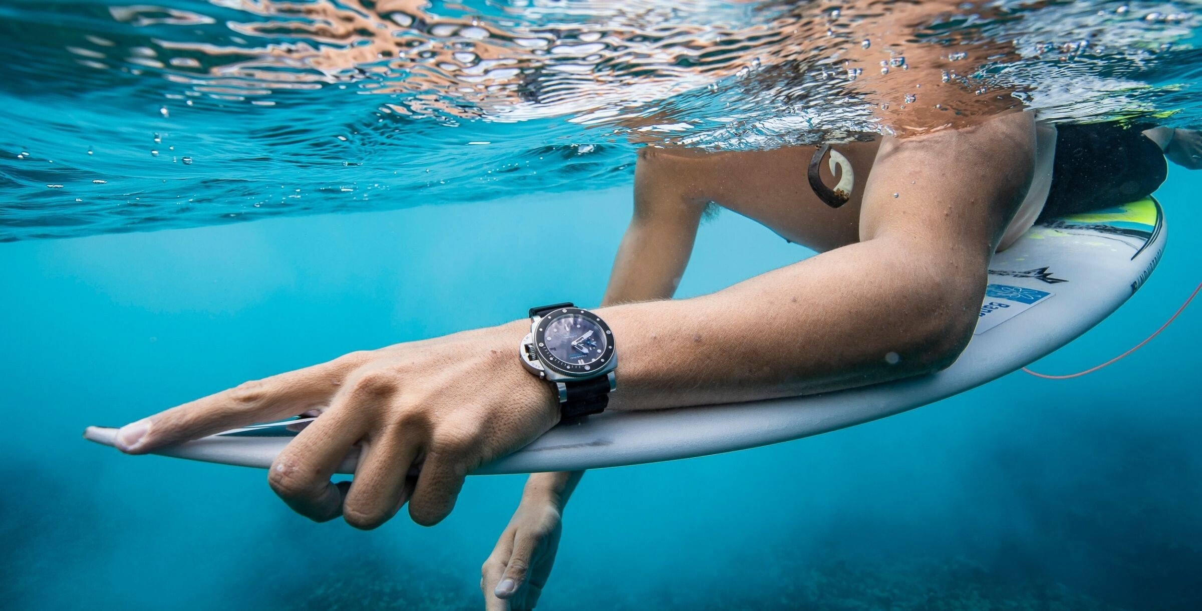 Imagem de um surfista parcialmente debaixo de água em cima da sua prancha e com um relógio Panerai no pulso em destaque