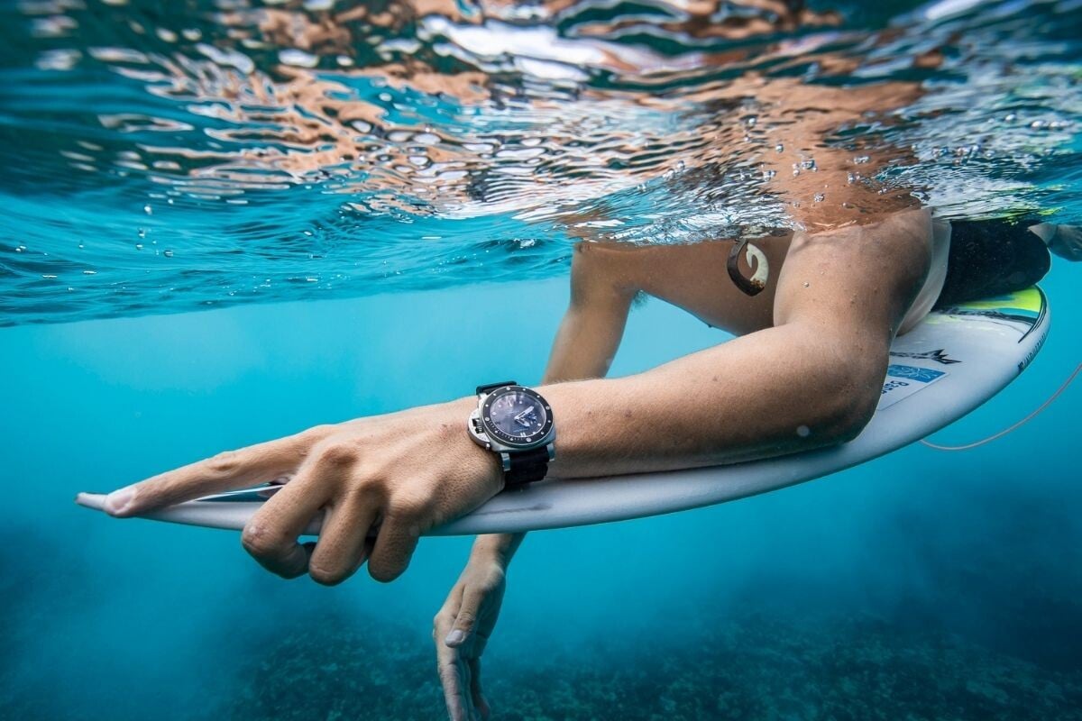 Imagem de um surfista parcialmente debaixo de água em cima da sua prancha e com um relógio Panerai no pulso em destaque