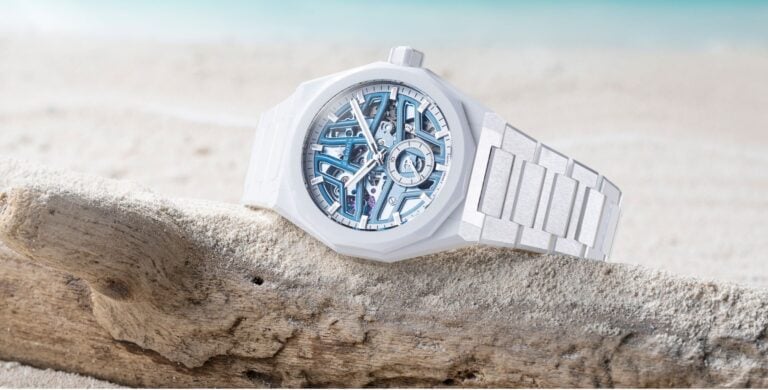 Relógio Zenith DEFY Skyline Skeleton White Ceramic pousado sobre um tronco de madeira na areia, com o mar desfocado ao fundo.
