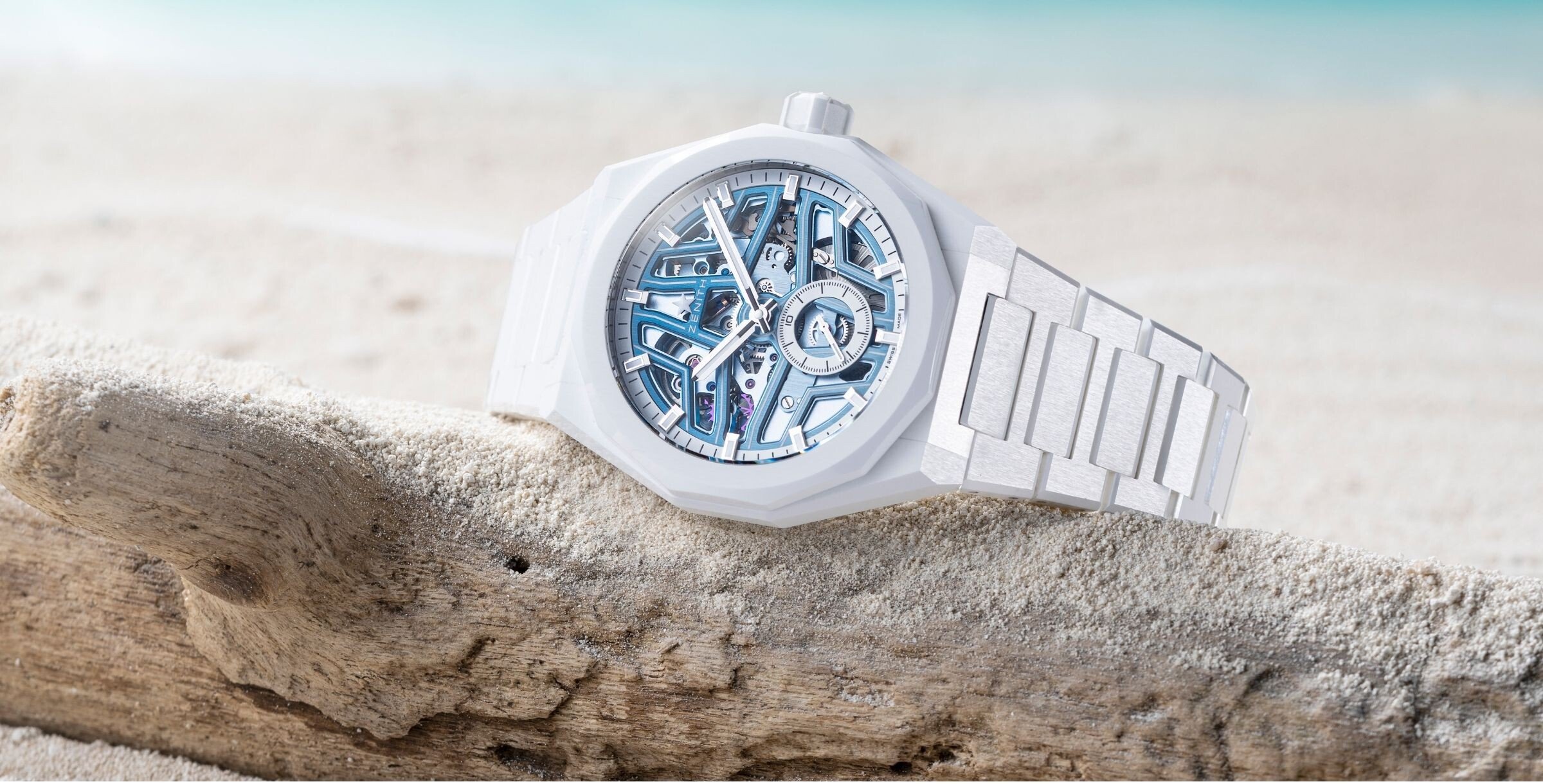 Relógio Zenith DEFY Skyline Skeleton White Ceramic pousado sobre um tronco de madeira na areia, com o mar desfocado ao fundo.