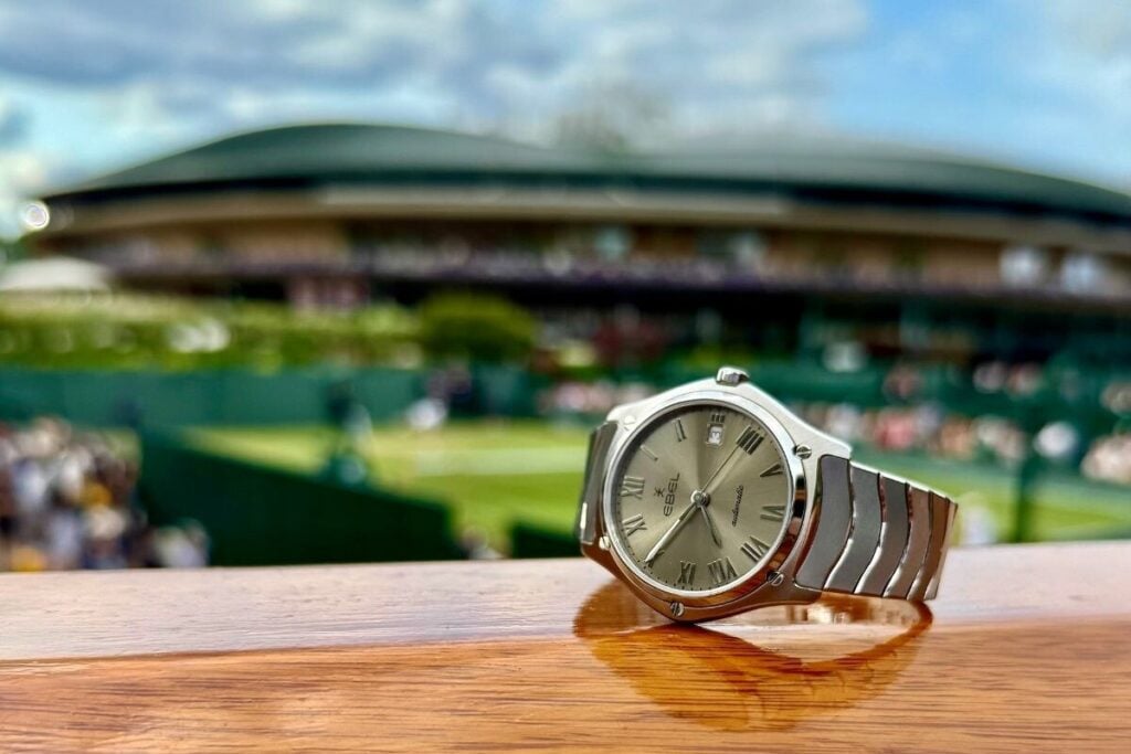 Relógio Ebel Classic Sport em primeiro plano sobre uma superfície de madeira, com um court de ténis de Wimbledon desfocado ao fundo.