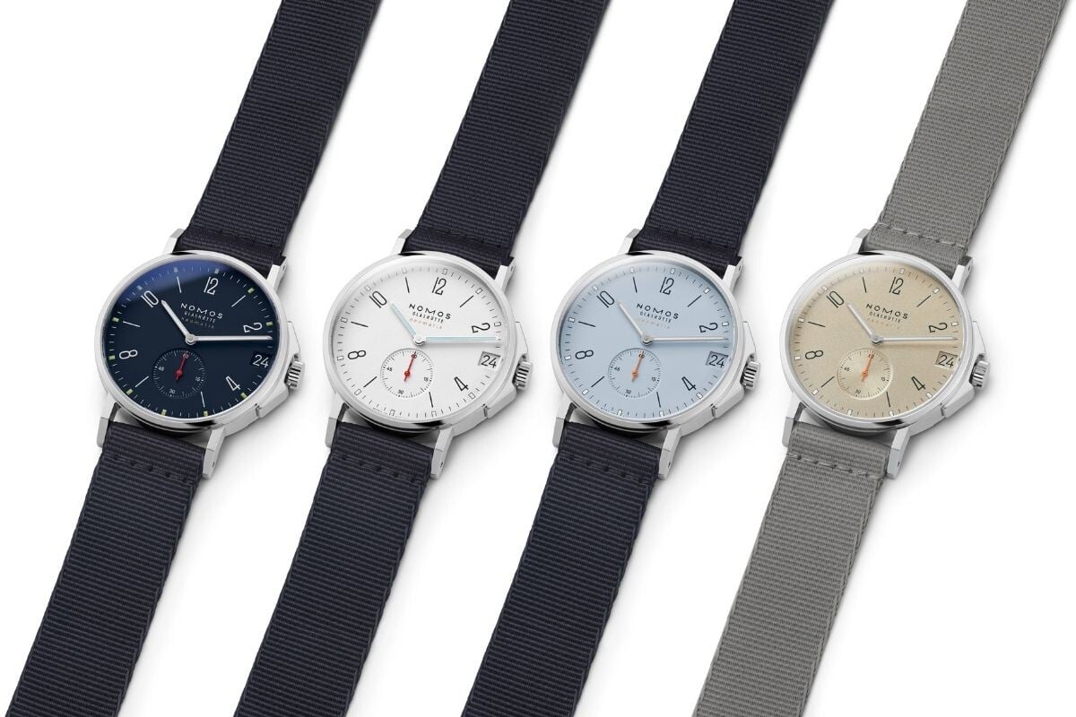 Quatro relógios de pulso da marca Nomos Glashütte alinhados lado a lado. Todos têm caixa de aço inoxidável e mostrador com design minimalista. Da esquerda para a direita: mostrador azul-escuro com pulseira azul-marinho, mostrador branco com pulseira azul-marinho, mostrador azul-claro com pulseira azul-marinho e mostrador bege-areia com pulseira cinza. Todos possuem pequenos segundos às 6 horas e janela de data às 3 horas.