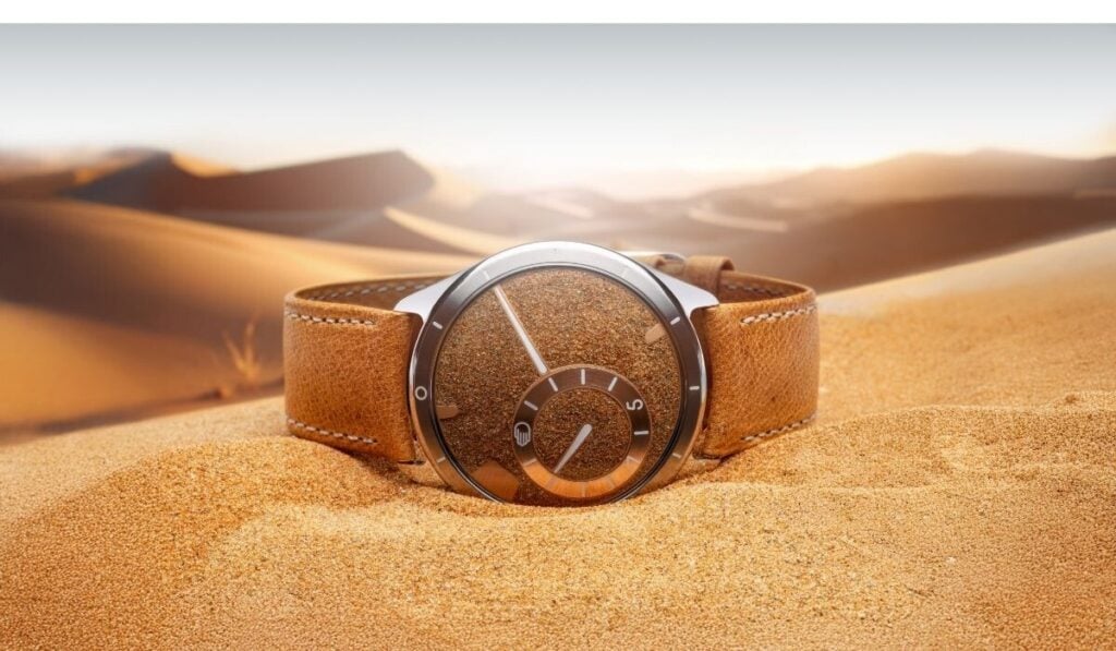 Relógio de pulso da marca Ressence, em titânio com mostrador preenchido de areia, deitado na areia, posição horizontal, onde se vislumbram ao longe as dunas.