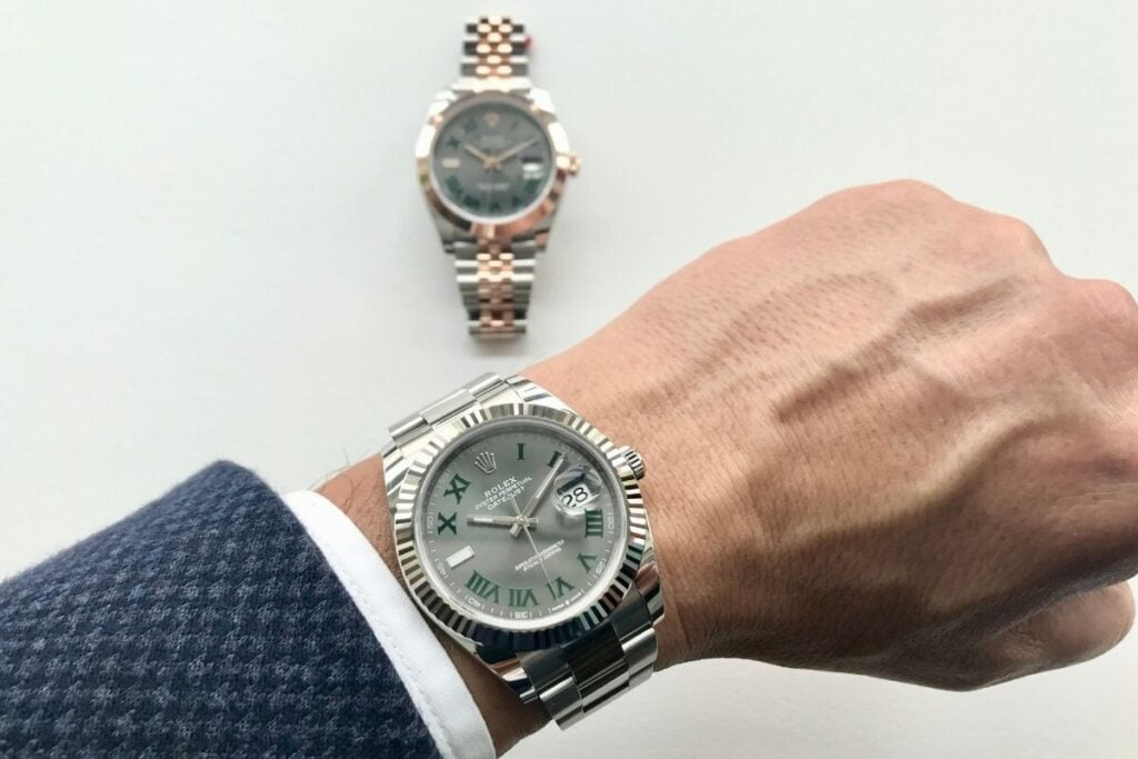Pessoa com relógio Rolex Datejust no pulso, com outro modelo bicolor em segundo plano sobre superfície branca.