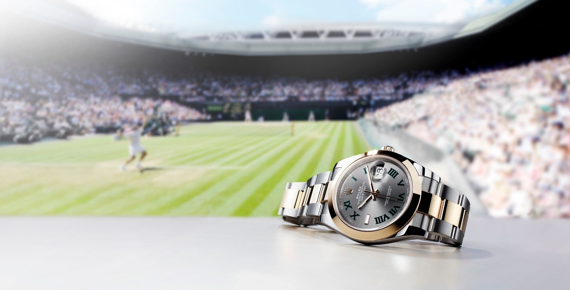 Relógio Rolex Datejust com mostrador wimbledon cinzento fotografado em primeiro plano, com uma partida de ténis ao fundo em estádio lotado.