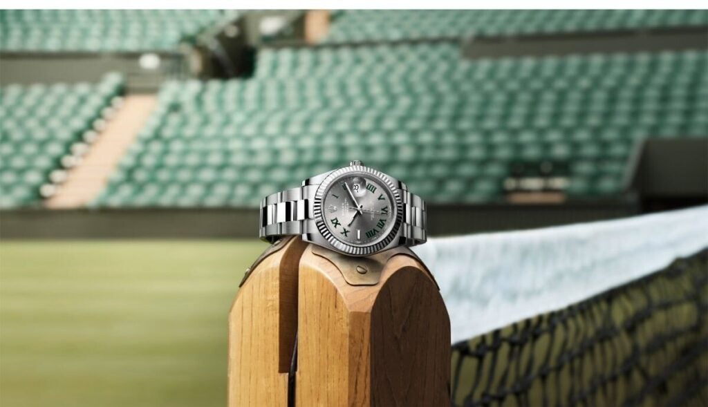 Relógio Rolex Datejust com mostrador cinza, algarismos romanos verdes e bracelete Oyster em primeiro plano e ao fundo a plateia do court
