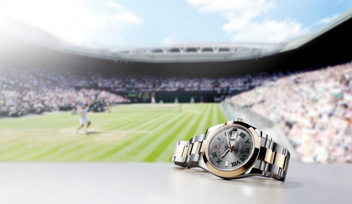 Relógio Rolex Datejust com mostrador wimbledon cinzento fotografado em primeiro plano, com uma partida de ténis ao fundo em estádio lotado.