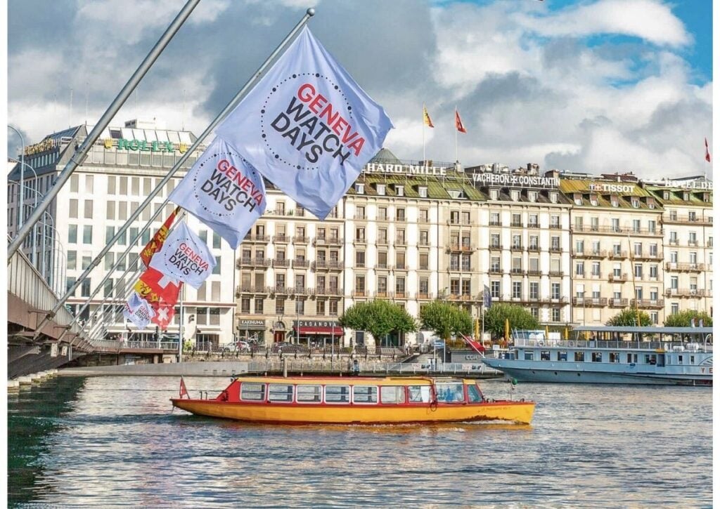 Bandeiras do Geneva Watch Days hasteadas em ponte sobre o Lago Léman, com edifícios históricos e barco turístico ao fundo.