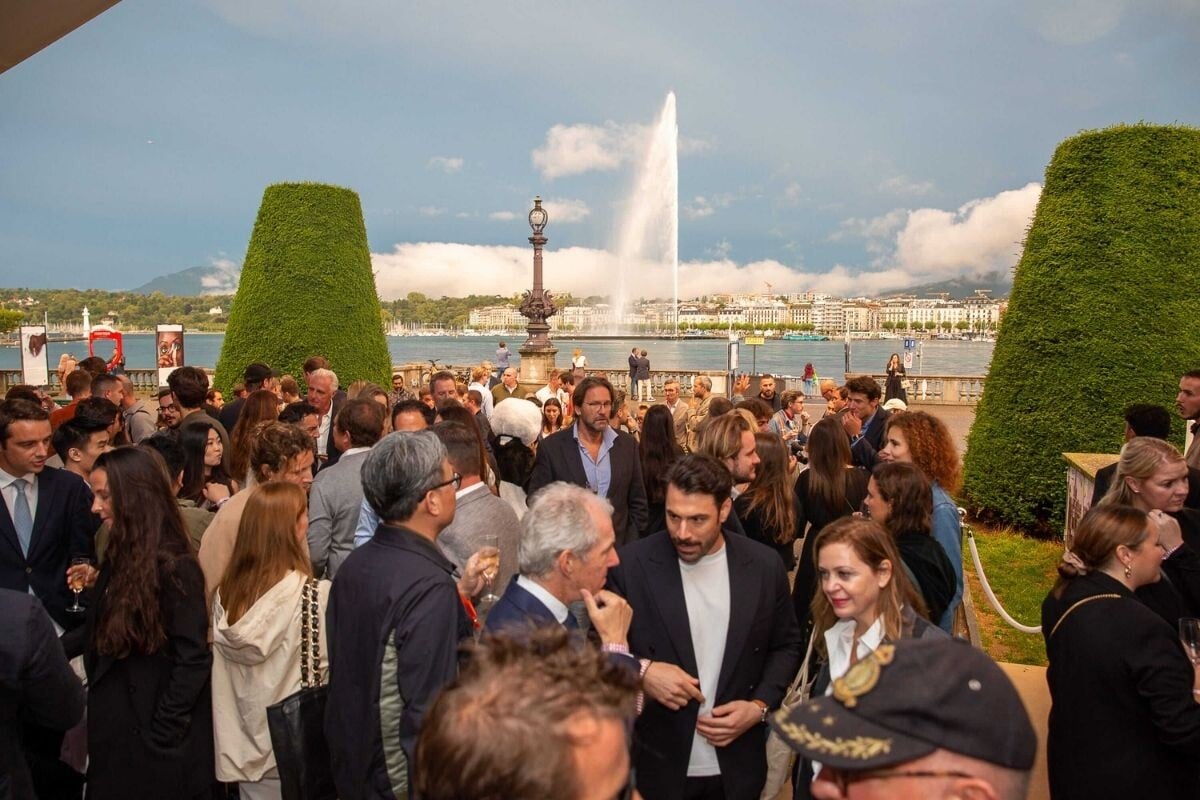 Pessoas reunidas ao ar livre durante o Geneva Watch Days, com o Jet d’Eau iluminado ao fundo ao entardecer.