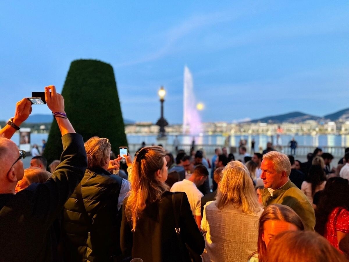 Pessoas reunidas ao ar livre durante o Geneva Watch Days, com o Jet d’Eau iluminado ao fundo ao entardecer.