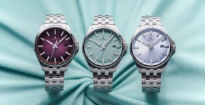 Três relógios Junghans de pulso em aço inoxidável com mostradores em tons de borgonha, verde-água e azul-claro, alinhados lado a lado sobre um fundo acetinado azul-turquesa.