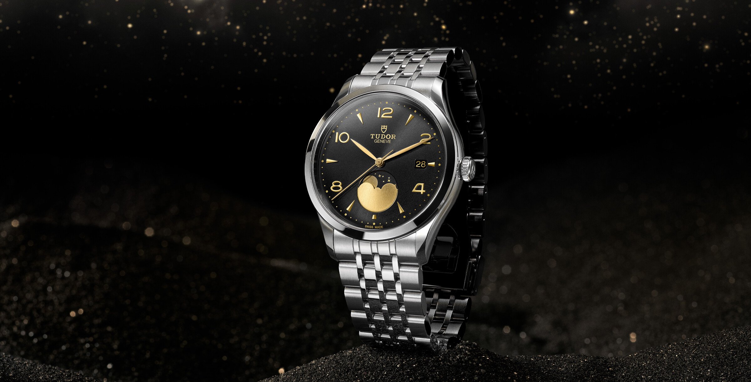 Relógio Tudor 1926 Moonphase em aço com mostrador preto e detalhes dourados, fotografado sobre fundo escuro estrelado.