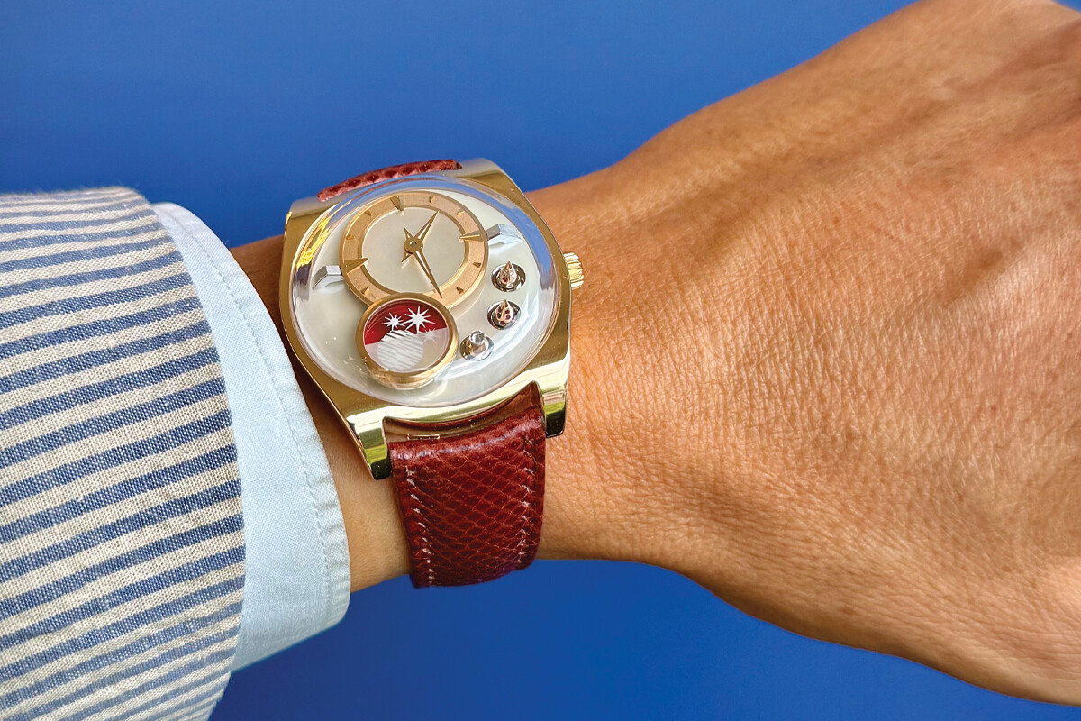 Relógio de pulso elegante com caixa dourada, mostrador claro e pulseira de couro vermelho, usado no pulso de uma pessoa vestindo camisa listrada azul e branca.