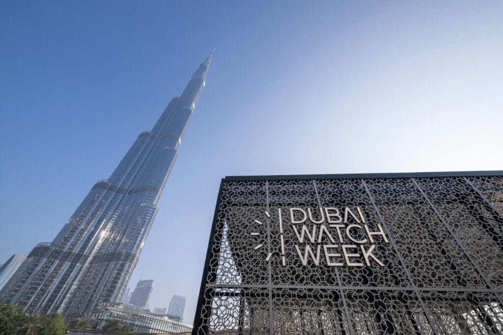 A imagem mostra a entrada do evento Dubai Watch Week, com o icônico Burj Khalifa ao fundo. A entrada é uma estrutura moderna com um design intrincado, exibindo o nome "DUBAI WATCH WEEK" em letras grandes e elegantes.