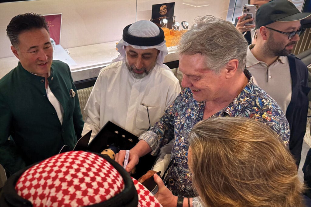 Pessoas interagindo em uma loja de relógios de luxo durante a Dubai Watch Week. Um homem está segurando um relógio dentro de uma caixa enquanto outros observam com interesse. A cena mostra uma mistura de pessoas vestindo roupas formais e tradicionais árabes, com um ambiente sofisticado ao fundo, com exibição de relógios.
