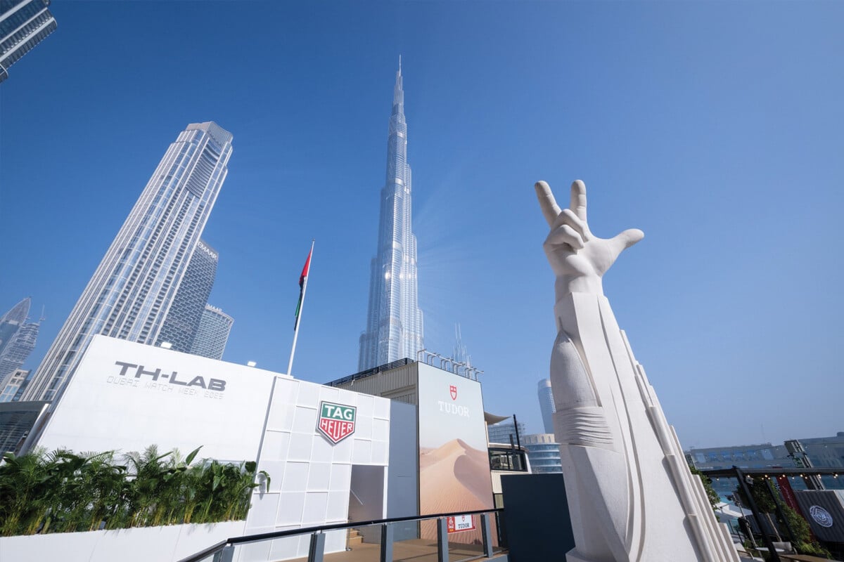 Vista do Burj Khalifa ao fundo, com instalações da Dubai Watch Week em primeiro plano, incluindo um pavilhão da TAG Heuer e uma grande escultura branca de uma mão fazendo um gesto com três dedos levantados.