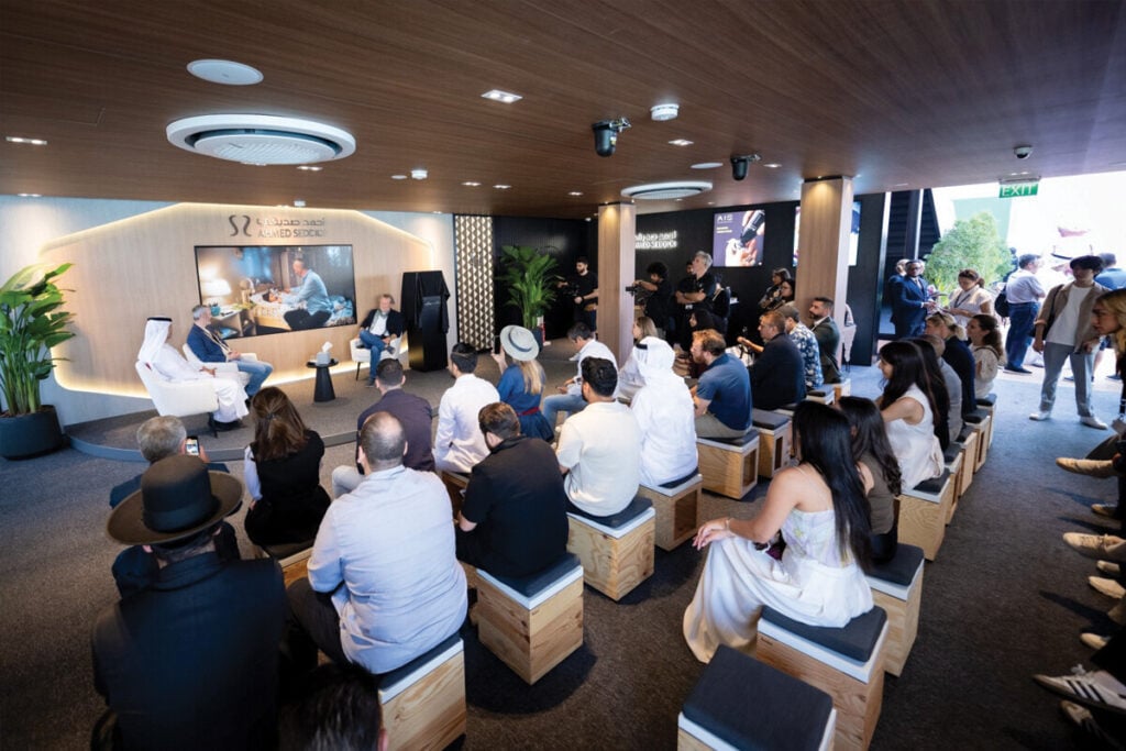 Um evento na Dubai Watch Week com uma apresentação acontecendo no palco. O público está sentado em cadeiras baixas de madeira, assistindo aos palestrantes. Há um grande painel de fundo com uma imagem e dois oradores no palco. Pessoas estão filmando e tirando fotos, enquanto a sala é bem iluminada e moderna.