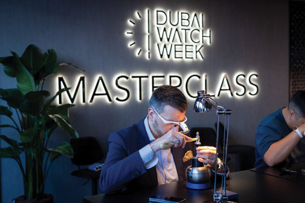 Um homem participa de uma masterclass na Dubai Watch Week, trabalhando em um relógio com ferramentas precisas. Ele usa óculos de aumento e está concentrado no trabalho. Ao fundo, uma luz neon exibe a palavra "MASTERCLASS", e há uma planta ao lado.