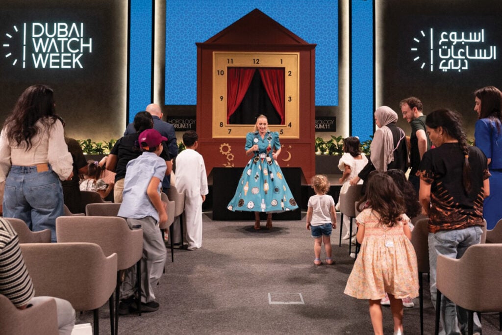 Uma apresentação interativa durante a Dubai Watch Week, com uma mulher vestida com um vestido azul estampado com relógios, interagindo com um grupo de crianças e adultos. O cenário inclui uma grande caixa de relógio com números e uma cortina vermelha. Pessoas estão saindo de seus assentos para participar da atividade.