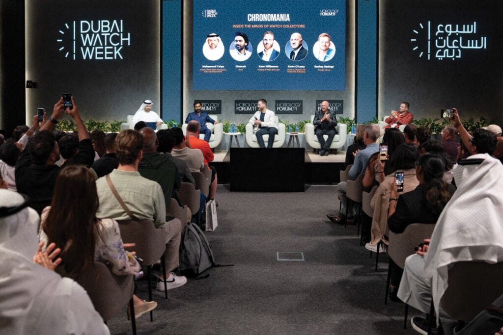 Público e palestrantes no evento Dubai Watch Week. No palco, um painel de discussão com cinco participantes, e o público na frente está tirando fotos e prestando atenção.