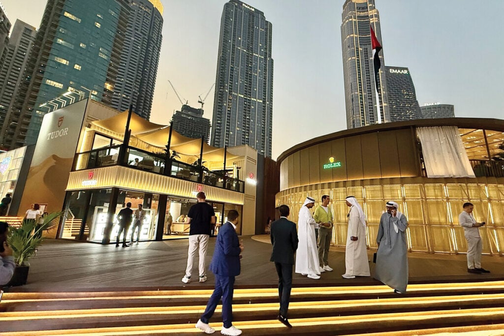 Uma cena noturna na Dubai Watch Week, mostrando lojas luxuosas como Tudor e Rolex. Há várias pessoas na área, algumas conversando e outras caminhando, enquanto edifícios altos de Dubai iluminam o fundo. Pessoas vestidas em trajes formais e tradicionais árabes estão interagindo com outras pessoas vestindo roupas mais casuais. O ambiente moderno é iluminado com luzes quentes.