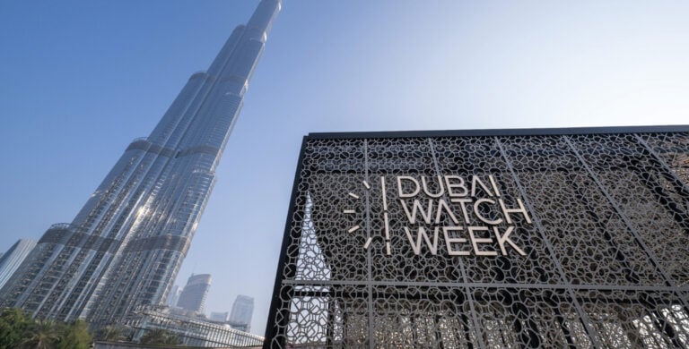 A imagem mostra a entrada do evento Dubai Watch Week, com o icônico Burj Khalifa ao fundo. A entrada é uma estrutura moderna com um design intrincado, exibindo o nome "DUBAI WATCH WEEK" em letras grandes e elegantes.