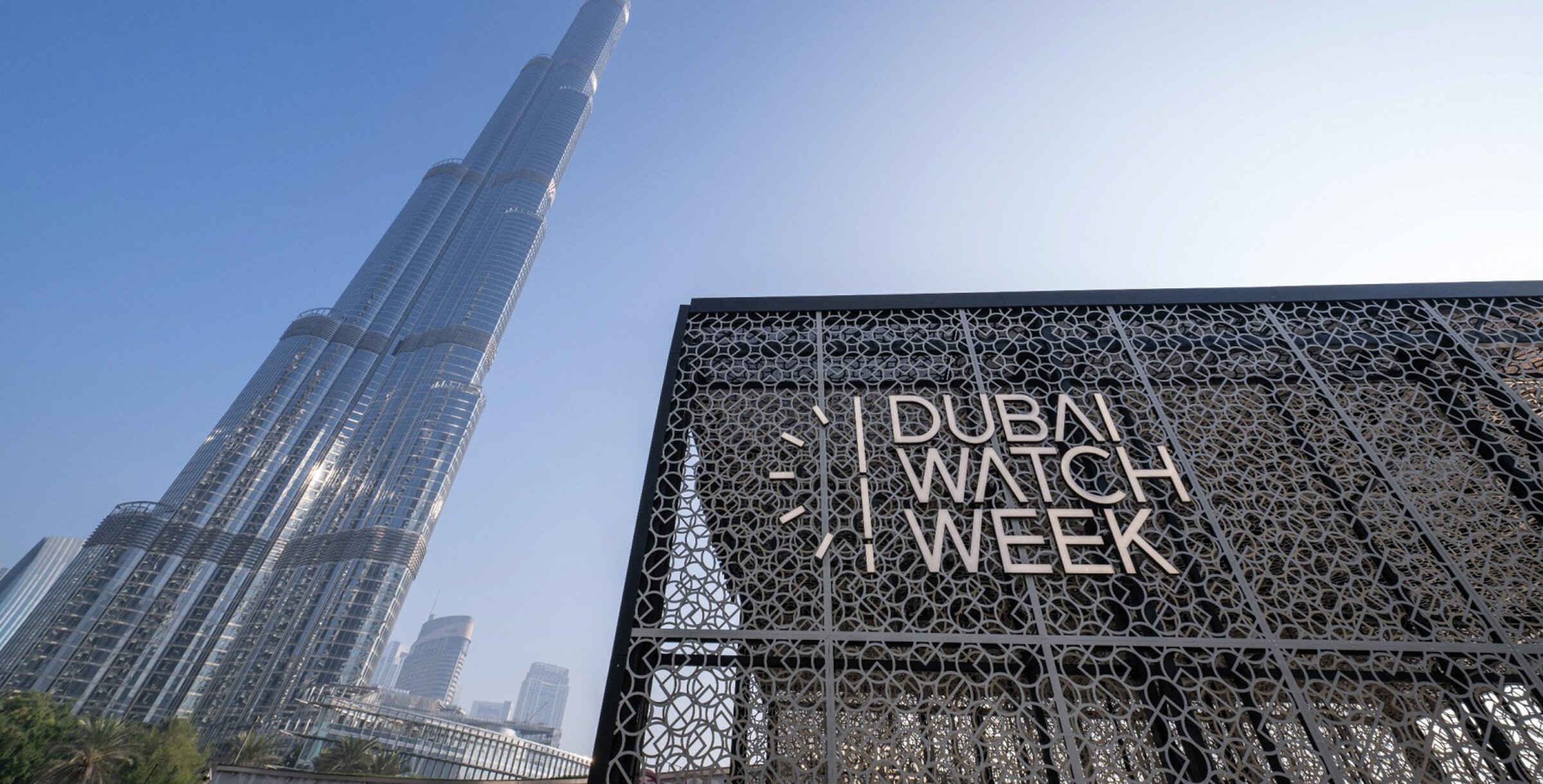 A imagem mostra a entrada do evento Dubai Watch Week, com o icônico Burj Khalifa ao fundo. A entrada é uma estrutura moderna com um design intrincado, exibindo o nome "DUBAI WATCH WEEK" em letras grandes e elegantes.