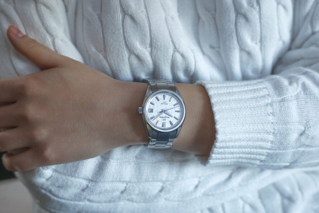 Pessoa a usar um relógio de pulso em aço prateado com mostrador branco, vestindo uma camisola de malha branca.