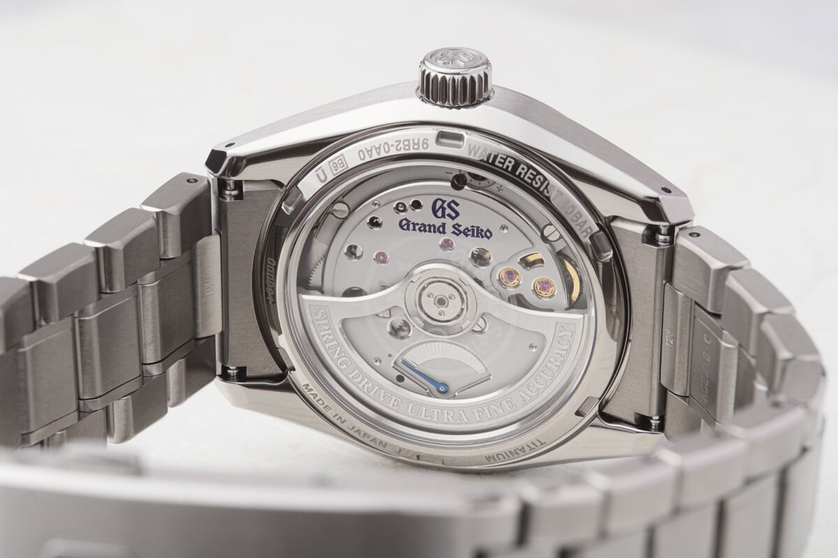 Vista traseira de um relógio de pulso metálico, mostrando o movimento mecânico visível através do fundo em vidro.