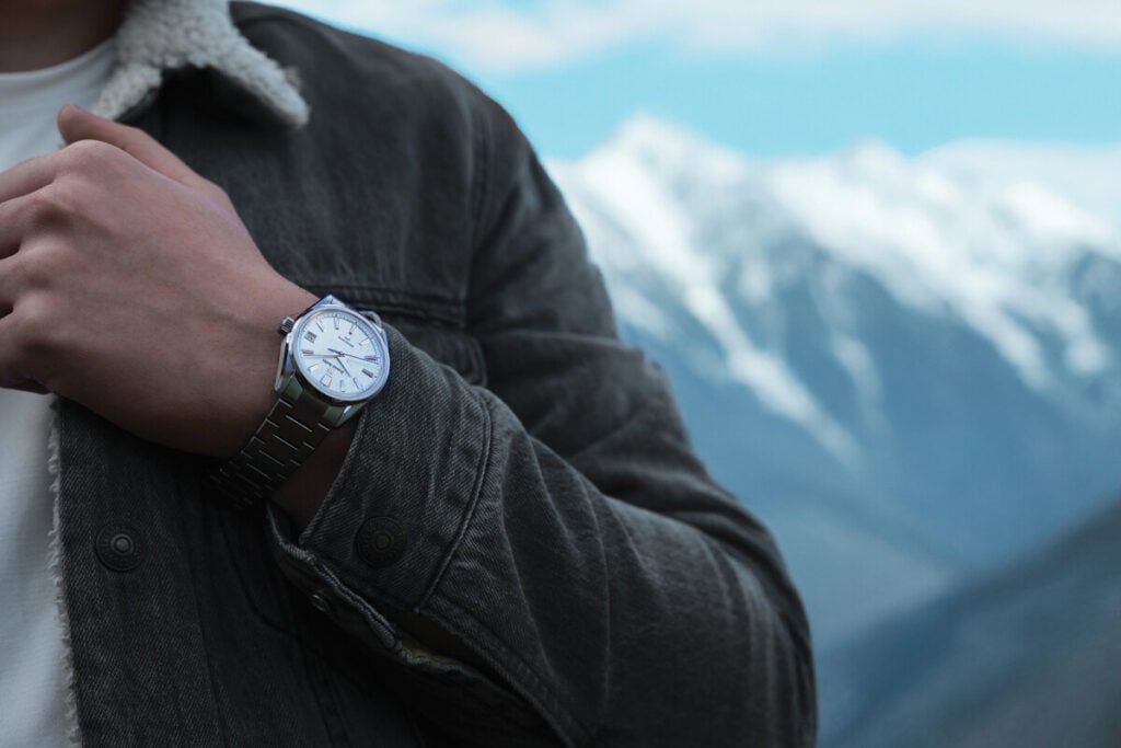 Pessoa a usar um relógio de pulso prateado com mostrador branco, em frente a uma paisagem de montanhas cobertas de neve.