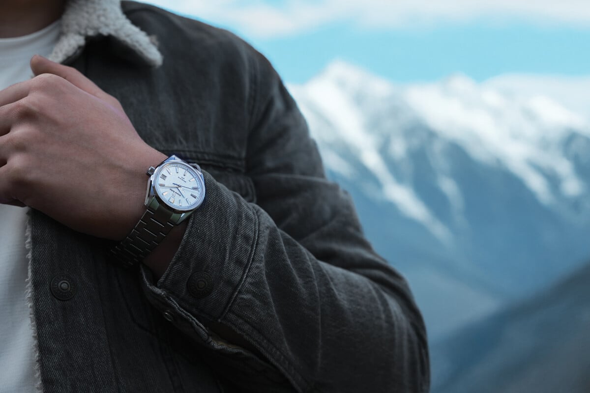 Pessoa a usar um relógio de pulso prateado com mostrador branco, em frente a uma paisagem de montanhas cobertas de neve.