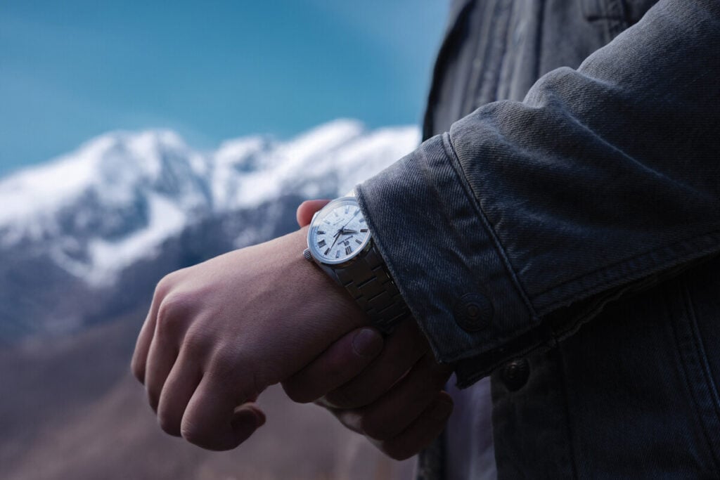 Pessoa a ajustar um relógio de pulso prateado com mostrador branco, com montanhas nevadas ao fundo.