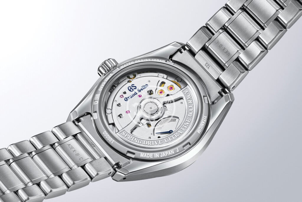 Vista traseira de um relógio de pulso metálico, mostrando o movimento mecânico visível através do fundo em vidro.