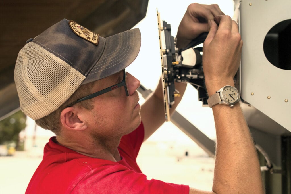"Homem usando óculos e boné, ajustando um equipamento com as mãos, enquanto usa um relógio de pulso com uma bracelete de tecido cinza. O fundo mostra o ambiente exterior com luz natural."