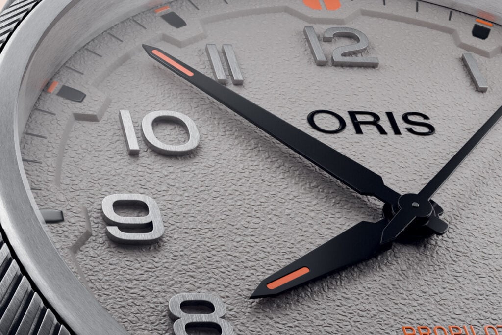 Imagem em close-up do mostrador de um relógio da marca Oris. É visível a textura detalhada do fundo do mostrador, com números grandes e finos em aço inoxidável. As horas estão indicadas com números arábicos, e as agulhas do relógio têm detalhes em preto e laranja, com a agulha dos minutos destacando-se pelo formato e pela cor. O logótipo "ORIS" está no centro, acima das 6 horas, e na parte inferior, próximo ao logo, está escrito "PRO PILOT".