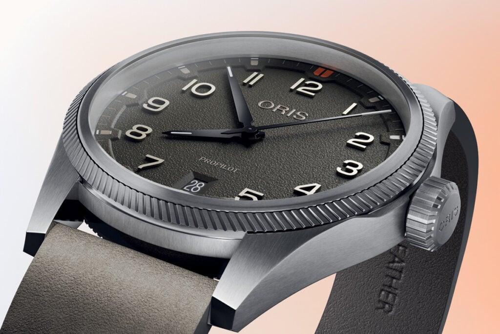 Imagem de close-up de um relógio Oris Pro Pilot com uma caixa de aço inoxidável e pulseira de couro. O mostrador é de cor verde escura, com números grandes e visíveis, e a data está na posição das 6 horas. O logótipo "ORIS" é exibido no topo do mostrador, e há um pequeno detalhe em laranja ao lado do número 12. O relógio tem uma caixa robusta, e a pulseira de couro é de cor clara, com um acabamento texturizado.