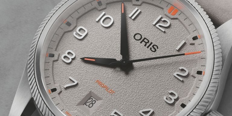 Relógio de pulso Oris ProPilot com mostrador cinza, num estilo moderno e robusto, com números e ponteiros de design limpo. O mostrador inclui a data num compartimento na posição das 6 horas, com detalhes em laranja. A caixa metálica escovada tem uma borda serrilhada em torno da coroa.