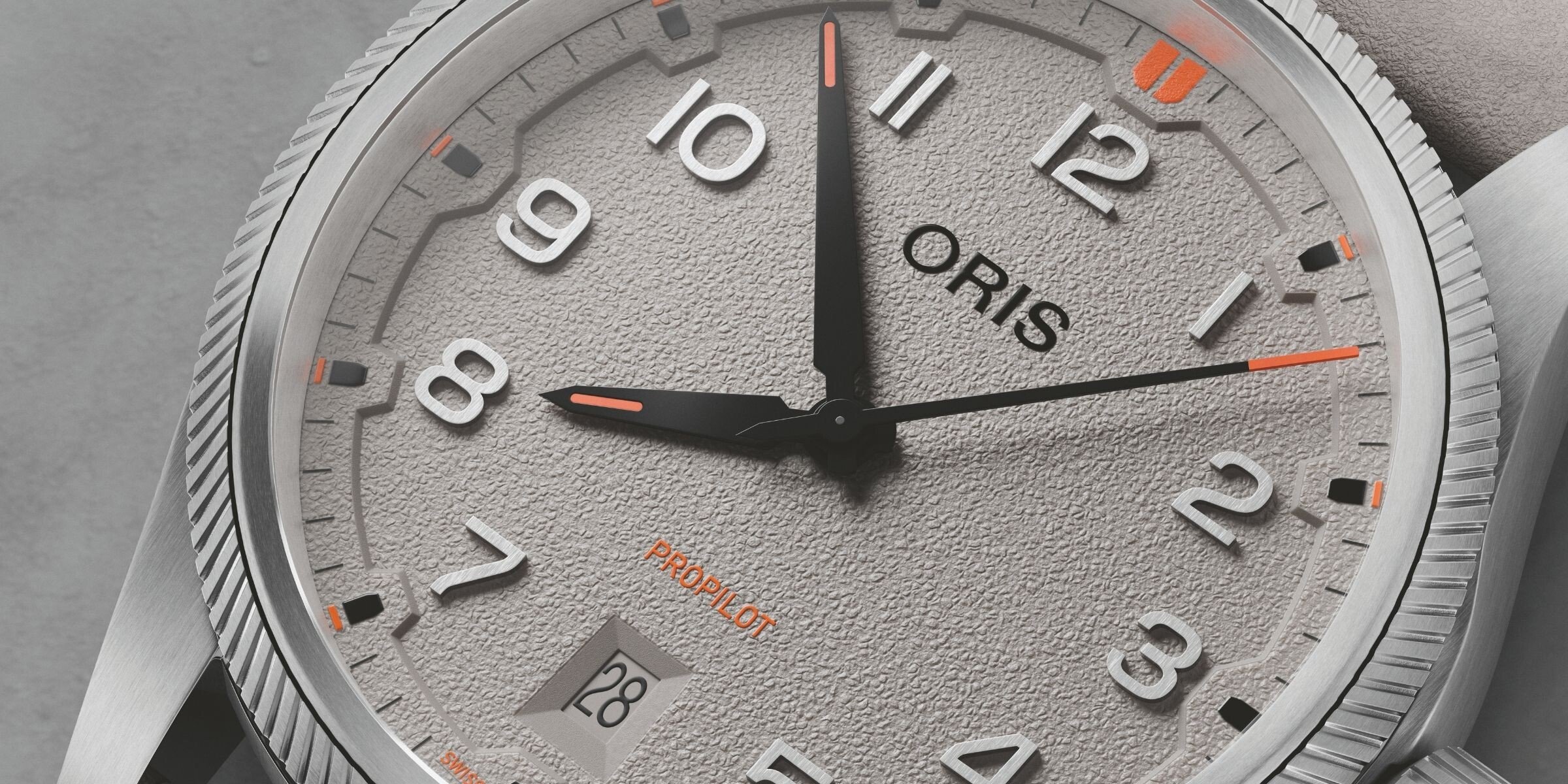 Relógio de pulso Oris ProPilot com mostrador cinza, num estilo moderno e robusto, com números e ponteiros de design limpo. O mostrador inclui a data num compartimento na posição das 6 horas, com detalhes em laranja. A caixa metálica escovada tem uma borda serrilhada em torno da coroa.