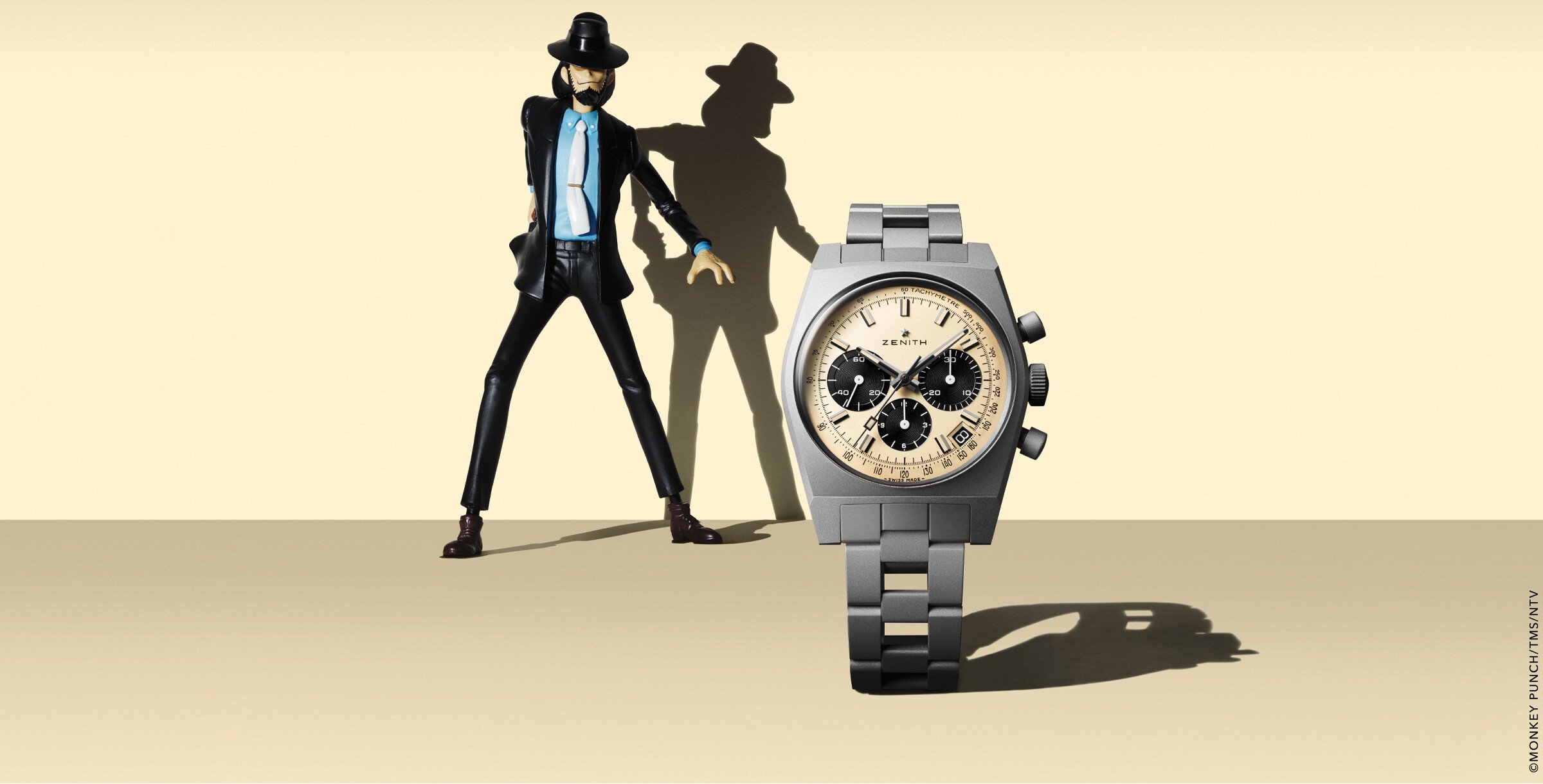 Imagem de um relógio Zenith Chronomaster Revival Daisuke Jigen Edition ao lado de uma figura de ação do personagem Daisuke Jigen, de "Lupin III". O relógio está em um fundo neutro, com o destaque para o design vintage da peça e a figura estilizada de Jigen projetando uma sombra longa no fundo.