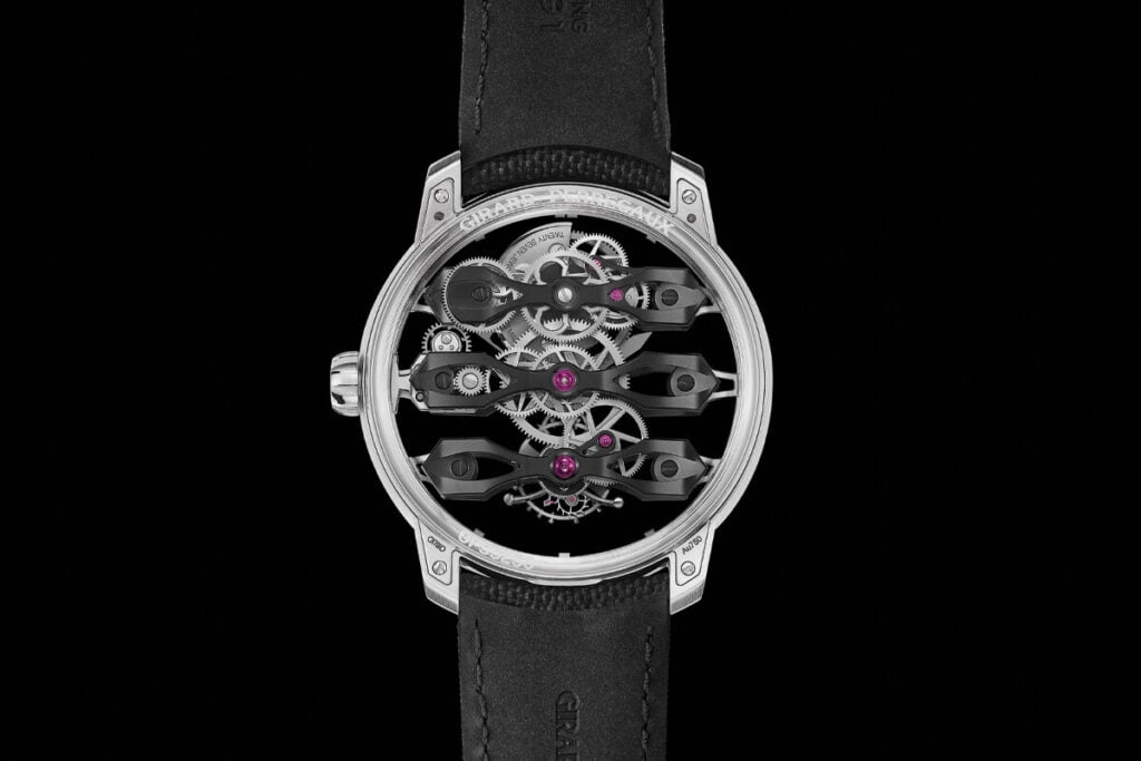 Rel&oacute;gio Girard-Perregaux com uma caixa met&aacute;lica prateada, destacando um design transparente que revela seus complexos mecanismos internos. O mostrador apresenta tr&ecirc;s ponteiros flutuantes, vis&iacute;veis atrav&eacute;s da estrutura aberta, com v&aacute;rias pe&ccedil;as em metal escuro e rubis, visibilizando o tourbillon e os componentes de precis&atilde;o.