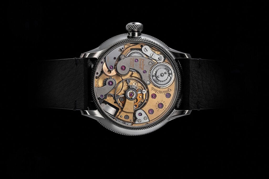 Vista traseira de um relógio mecânico com o movimento à vista, mostrando engrenagens douradas, rubis e componentes de precisão, preso a uma bracelete de couro preta sobre fundo escuro. Norifumi Seki (Quiet Club Fading Hours)