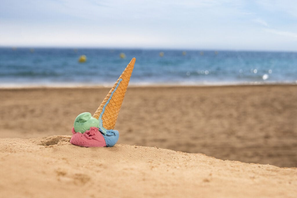 Imagem de um gelado de bolacha com bolas de gelado coloridas (verde, rosa e azul) parcialmente derretido, colocado na areia da praia, com o mar ao fundo.