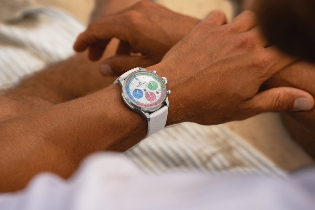 No pulso: rel&oacute;gio autom&aacute;tico Norqain Freedom 60 40mm Enjoy Life Special Edition em a&ccedil;o inoxid&aacute;vel, com cron&oacute;grafo e mostrador branco com submostradores coloridos em azul, verde e rosa, gelado na data com bracelete branca na praia