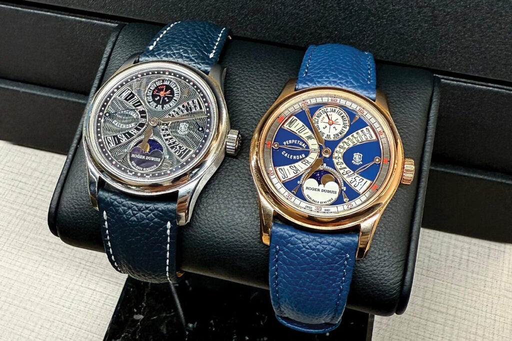Dois rel&oacute;gios de luxo da marca Roger Dubuis, ambos com pulseira de couro azul e mostradores detalhados, exibidos lado a lado em um suporte preto.