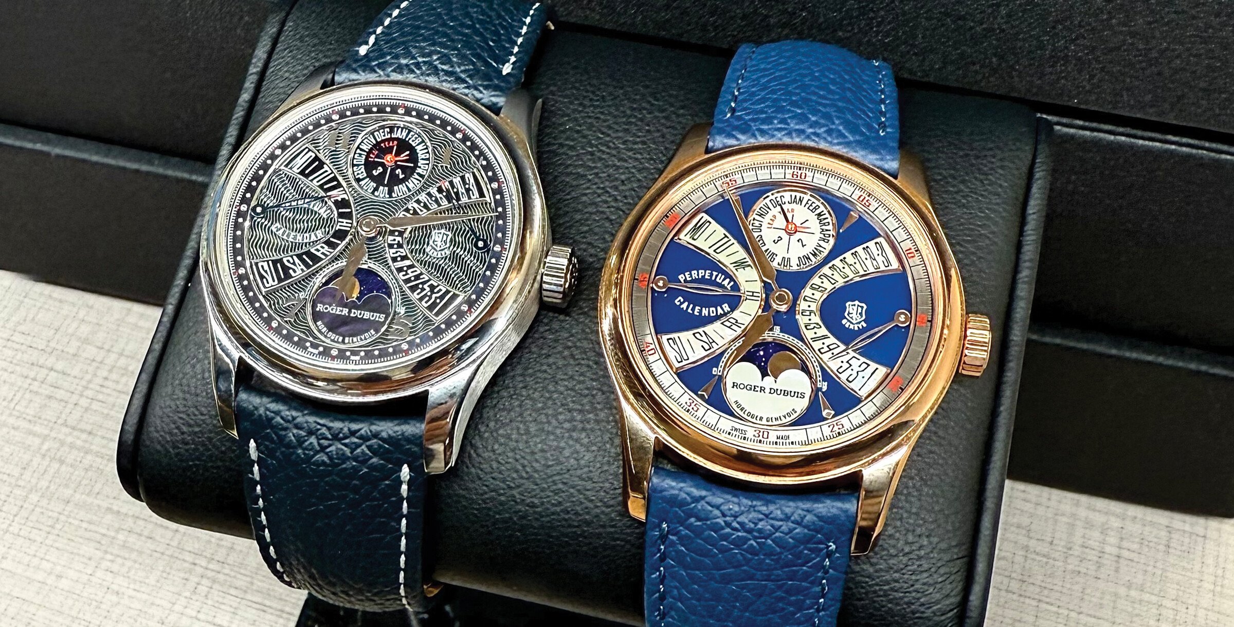 Dois relógios de luxo da marca Roger Dubuis, ambos com pulseira de couro azul e mostradores detalhados, exibidos lado a lado em um suporte preto.