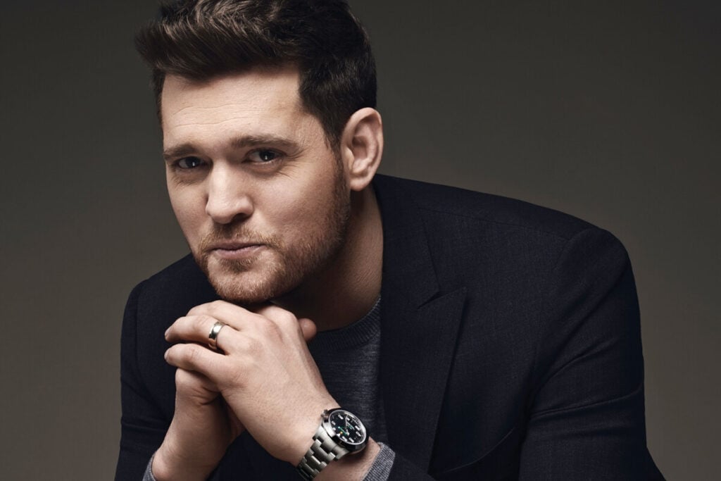 Homem com cabelo castanho curto e barba, vestindo um fato escuro e camisa cinza, com um relógio de pulso e mãos entrelaçadas perto do rosto, sorrindo suavemente. Michael Bubblé Rolex